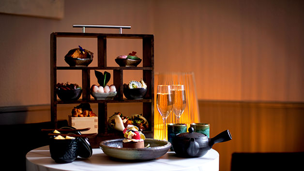 Contemporary afternoon tea presentation on round white table with dark wood box display stand holding savory items in black bowls, black teapot and cups, champagne flutes, and decorative plates with Asian-fusion dishes against warm ochre wall with ambient lighting