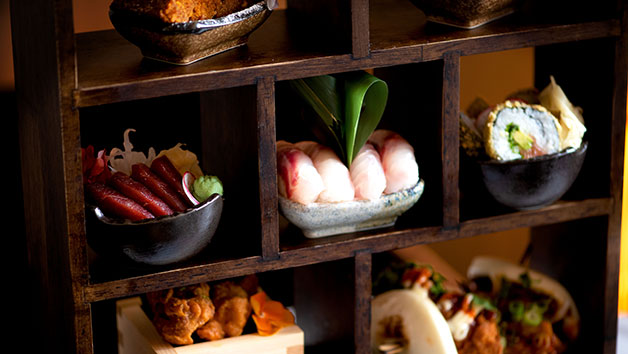 Dark wood cubby shelf display with Japanese-inspired afternoon tea items in black ceramic bowls and white dishes, featuring sushi rolls, pink nigiri with wasabi, tempura, colorful desserts, and various small plates arranged in compartments with dramatic lighting