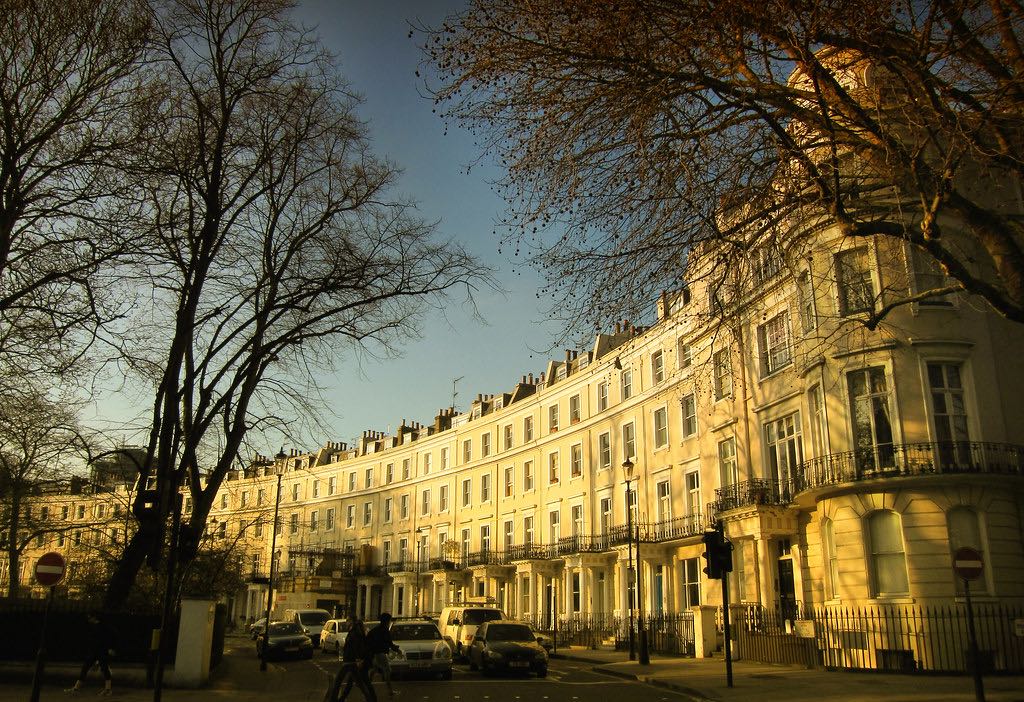 Georgian terraced townhouses in Bath with cream-colored stone facades glowing in golden evening light, multiple stories with uniform sash windows, iron balconies, street-level entrances with pillars, bare winter trees in foreground, parked cars along street, pedestrians walking